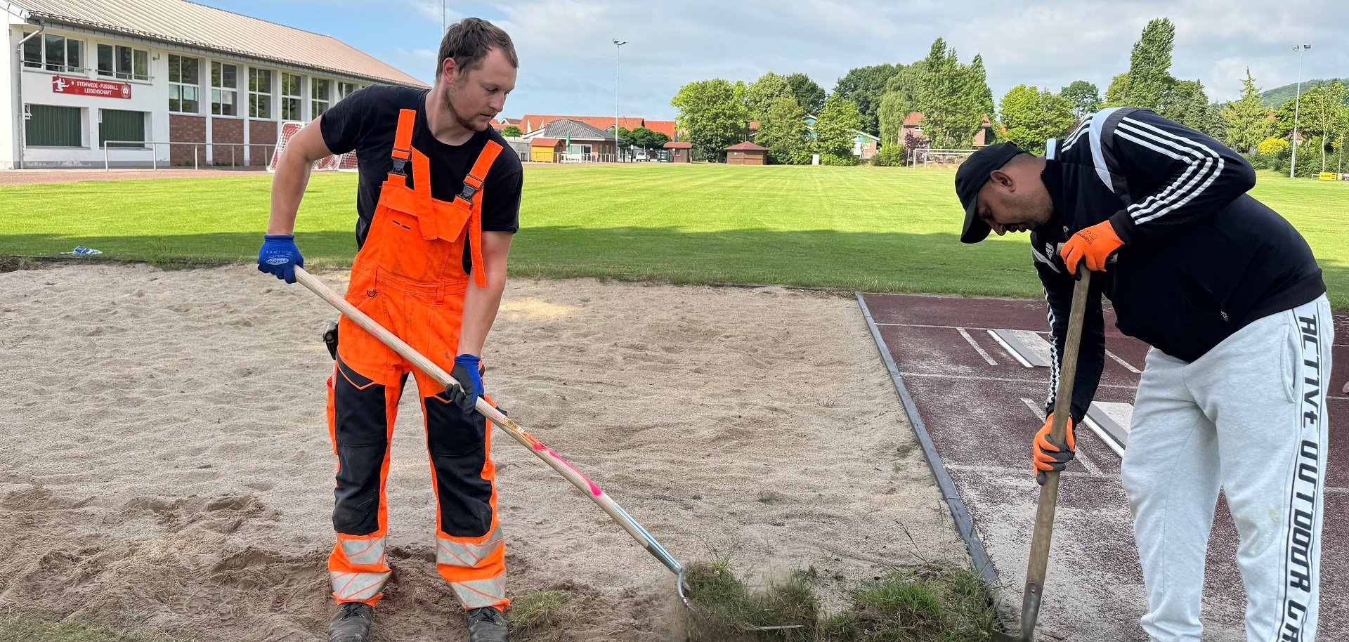 Zwei Männer arbeiten an einer Weitsprunggrube; einer trägt orange Arbeitskleidung und harkt Sand, der andere trägt Sportkleidung und gräbt am Rand mit einer Schaufel.