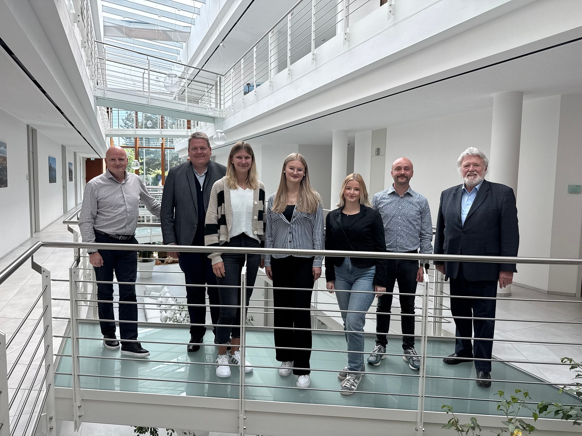 Insgesamt sieben Personen, vier Männer und drei junge Damen, haben sich in einem hellen Bürogebäude zum Gruppenfoto aufgestellt.