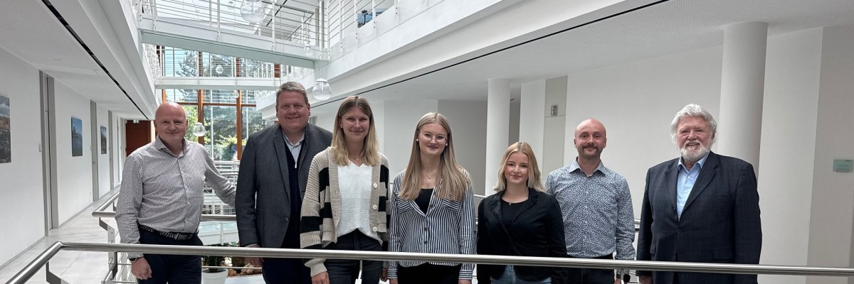 Insgesamt sieben Personen, vier Männer und drei junge Damen, haben sich in einem hellen Bürogebäude zum Gruppenfoto aufgestellt.