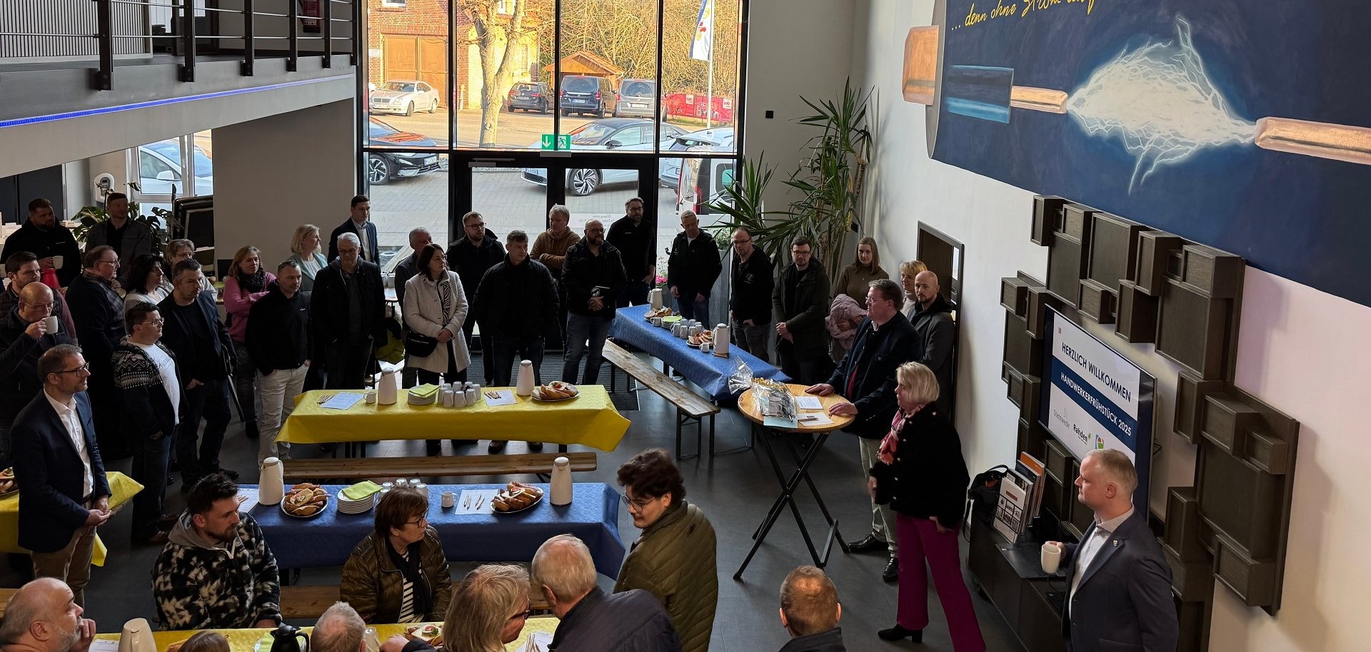Blick von oben ins Foyer eines Betriebes in dem mehrere Tische mit blauen und gelben Tischdecken aufgestellt sind an denen zahlreiche Personen sitzen und stehen.