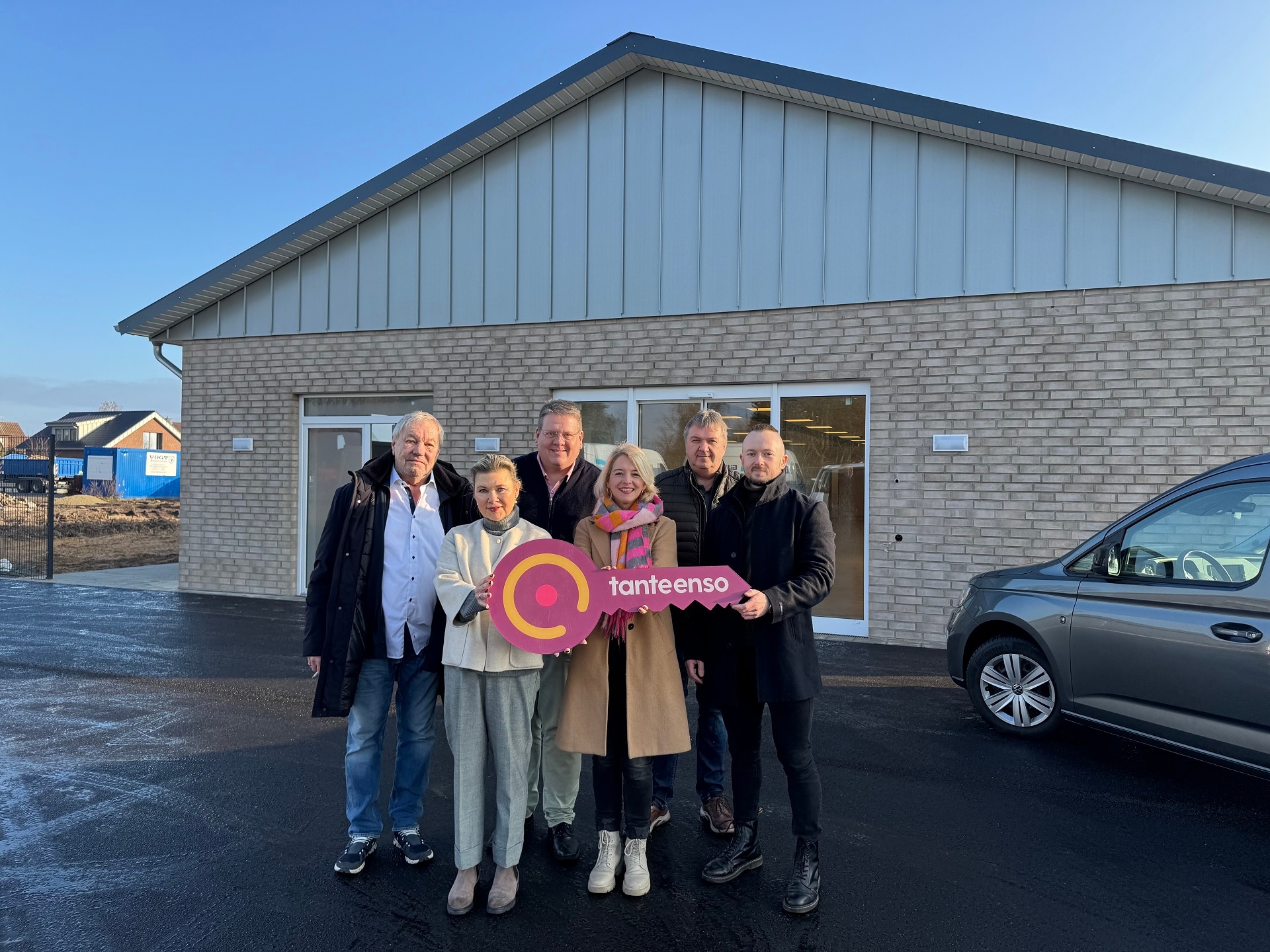 Eine Gruppe von Personen, zwei Frauen und vier Männer, die sich vor einem im Bau befindlichen Supermarktgebäude für ein Gruppenfoto aufgestellt haben. Die beiden vorne stehenden Frauen halten einen roten, überdimensionalen Schlüssel in der Hand auf dem "Tante Enso" steht.