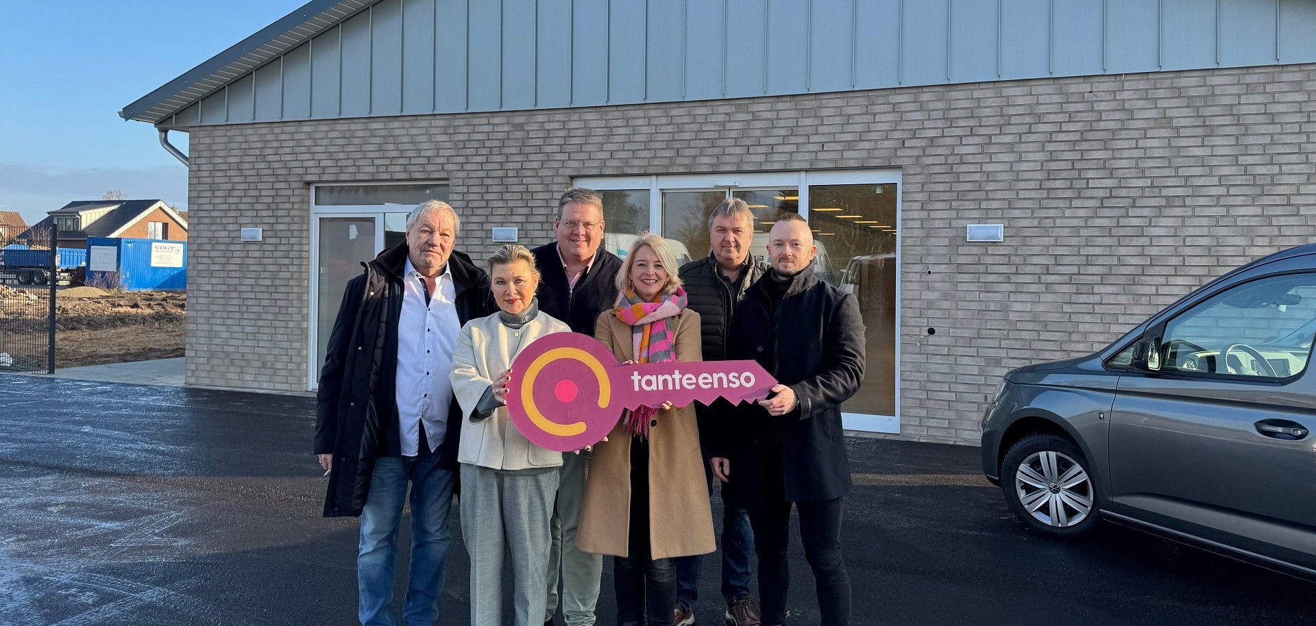 Eine Gruppe von Personen, zwei Frauen und vier Männer, die sich vor einem im Bau befindlichen Supermarktgebäude für ein Gruppenfoto aufgestellt haben. Die beiden vorne stehenden Frauen halten einen roten, überdimensionalen Schlüssel in der Hand auf dem "Tante Enso" steht.