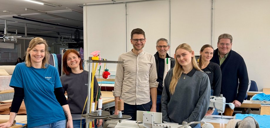 Sieben Personen stehen in einer hellen Polsterei-Werkstatt zwischen Arbeitstischen und Nähmaschinen und lächeln in die Kamera. Im Vordergrund befindet sich eine große Industrienähmaschine mit Garnrollen; daneben liegen Stoffe und Werkzeuge. Hinter der Gruppe sind weitere Näharbeitsplätze, Maßbänder und Polstermaterialien zu sehen. Einige tragen Arbeitskleidung mit Firmenlogo. Die Umgebung wirkt ordentlich und professionell.