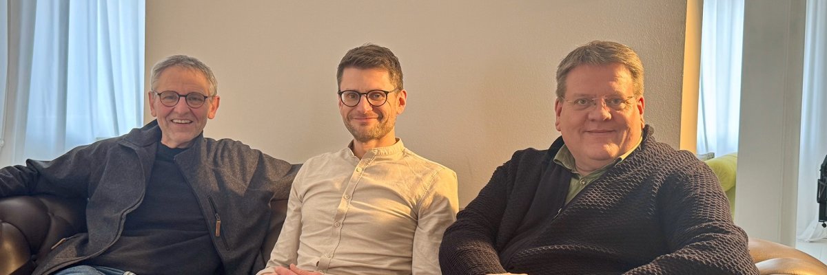 Drei Männer, die auf einem Ledersofa sitzen und in die Kamera lächeln.