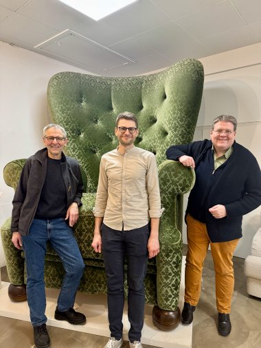 Drei Männer stehen in einem Innenraum vor einem sehr großen, grünen Ohrensessel mit hohem, geschwungenem Rücken und gemustertem Stoffbezug. Der Sessel ist deutlich überlebensgroß und wirkt wie eine überdimensionierte Version eines klassischen Polstersessels. Die Männer lächeln in die Kamera; einer steht mittig vor dem Sessel, die beiden anderen lehnen jeweils seitlich an den breiten Armlehnen. Der Raum ist hell beleuchtet, mit weißer Decke und neutralen Wänden.