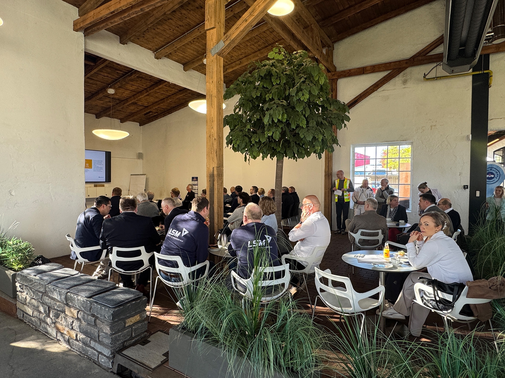 Handwerkerfrühstück 2026 Veranstaltung in einem großen Saal mit hoher Holzdecke und großen Fenstern. Zahlreiche Menschen sitzen an Tischen oder auf Stühlen und verfolgen einen Vortrag. Vorne stehen zwei Referenten neben einem Bildschirm mit Präsentationsfolie. In der Mitte des Raums steht ein großer grüner Innenbaum. Tageslicht erhellt den Raum, einige Teilnehmende stehen am Rand der Veranstaltung.