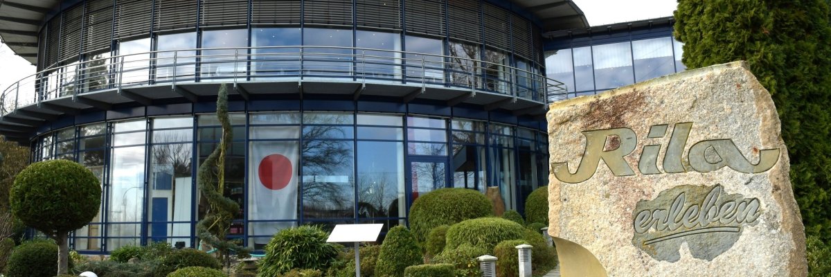 Modernes, rundes Gebäude mit großer Glasfassade und umlaufendem Balkon. Davor befindet sich ein gepflegter Garten mit geschnittenen Sträuchern, Steinwegen und kleinen Laternen. Im Vordergrund steht ein großer Stein mit der Inschrift „Rila erleben“. Durch die Fenster ist eine Dekoration mit einer japanischen Flagge zu sehen.