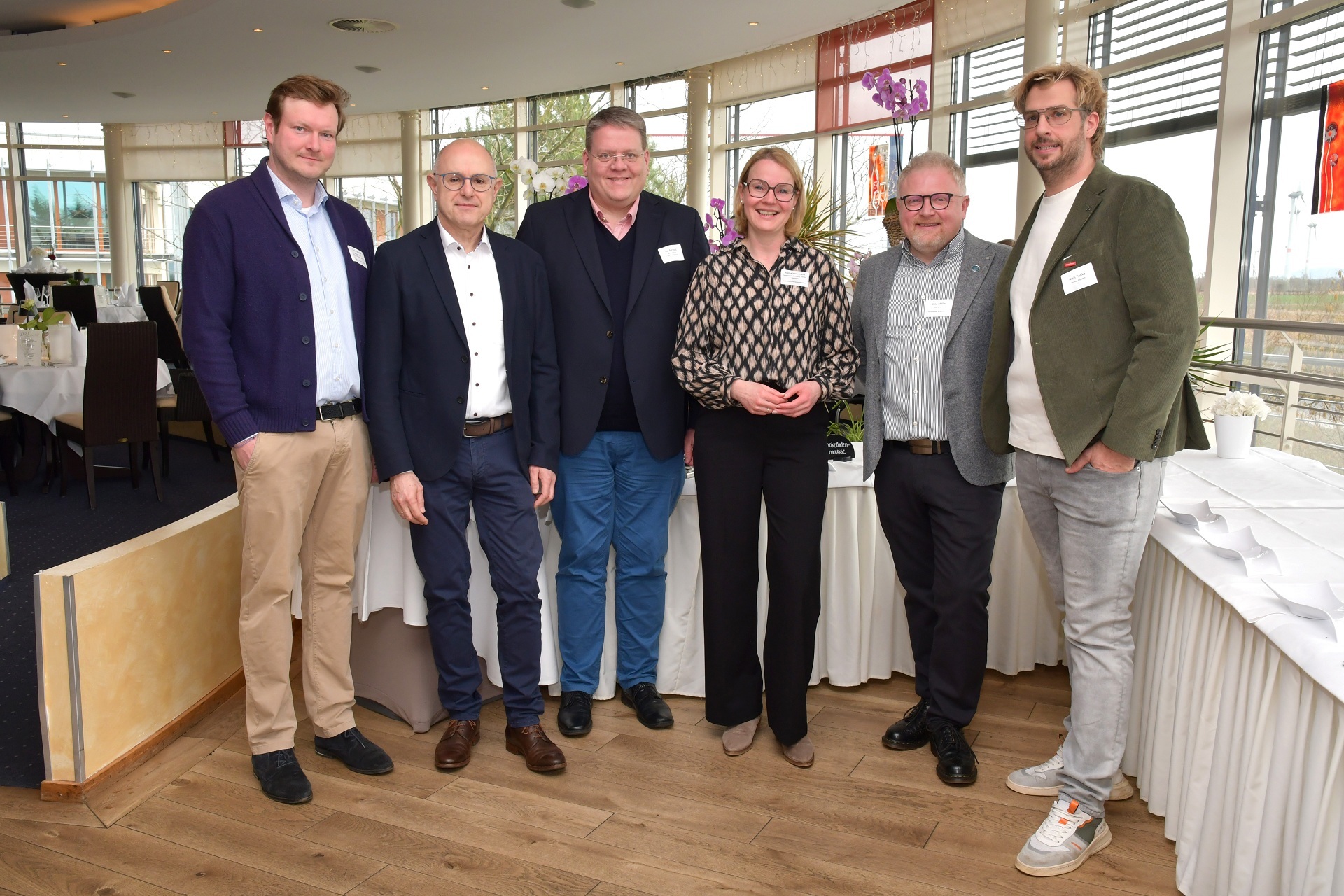 Sechs Erwachsene stehen nebeneinander in einem hellen Veranstaltungsraum mit großen Fenstern. Sie tragen Business- oder Business-Casual-Kleidung und Namensschilder und blicken in die Kamera. Im Hintergrund stehen runde, weiß gedeckte Tische mit Blumenarrangements; durch die Fenster ist eine ländliche Landschaft zu sehen. Die Gruppe wirkt wie Teil eines beruflichen Treffens oder Netzwerkevents.