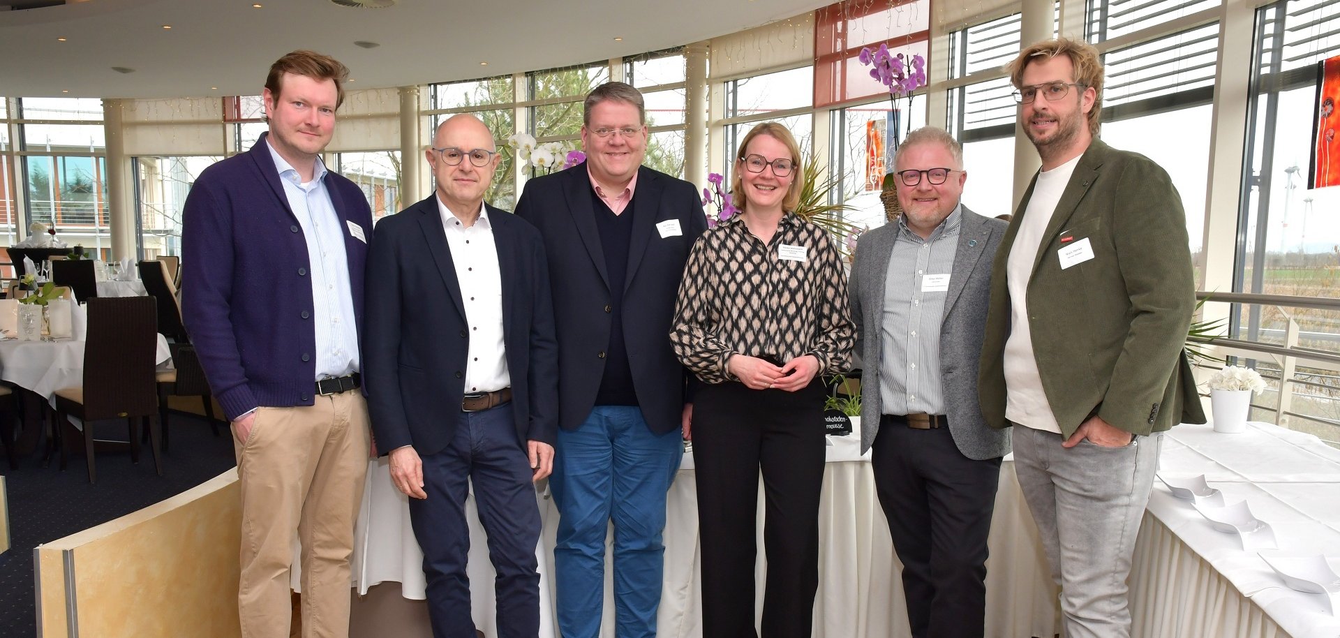 Sechs Erwachsene stehen nebeneinander in einem hellen Veranstaltungsraum mit großen Fenstern. Sie tragen Business- oder Business-Casual-Kleidung und Namensschilder und blicken in die Kamera. Im Hintergrund stehen runde, weiß gedeckte Tische mit Blumenarrangements; durch die Fenster ist eine ländliche Landschaft zu sehen. Die Gruppe wirkt wie Teil eines beruflichen Treffens oder Netzwerkevents.