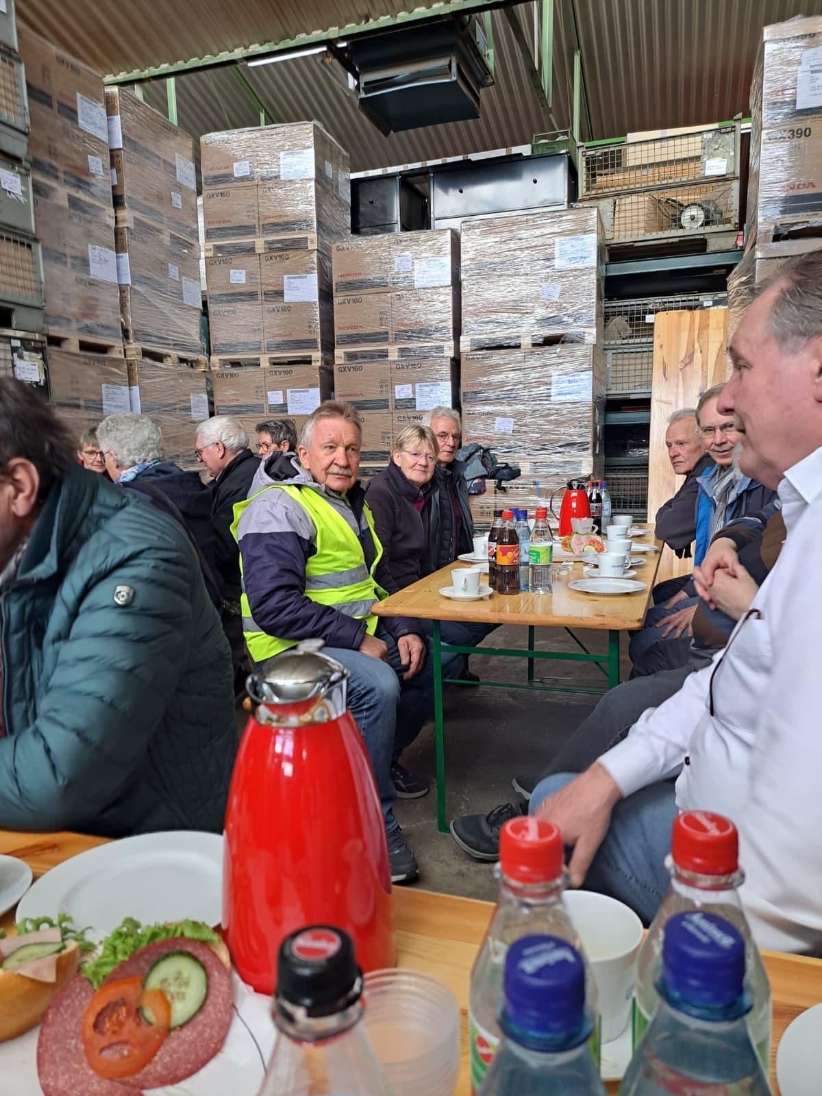 ZWAR-Tour Mehrere Menschen sitzen an langen Tischen in einer Lagerhalle zusammen und trinken Kaffee. Auf den Tischen stehen Getränke und belegte Brötchen. Im Hintergrund sind hohe Stapel palettierter Kartons und Regale zu sehen.