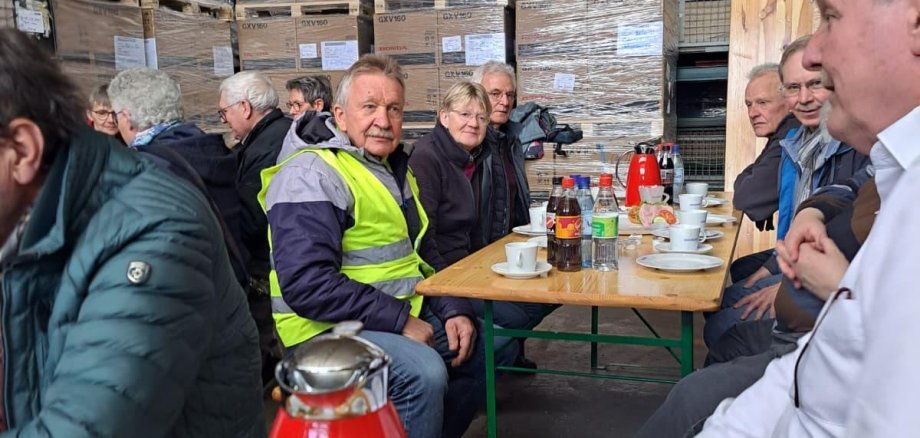 ZWAR-Tour Mehrere Menschen sitzen an langen Tischen in einer Lagerhalle zusammen und trinken Kaffee. Auf den Tischen stehen Getränke und belegte Brötchen. Im Hintergrund sind hohe Stapel palettierter Kartons und Regale zu sehen.