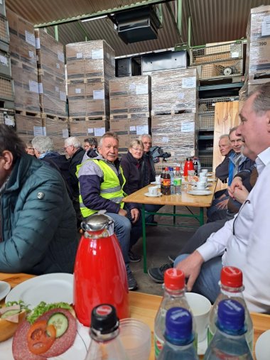 ZWAR-Tour Mehrere Menschen sitzen an langen Tischen in einer Lagerhalle zusammen und trinken Kaffee. Auf den Tischen stehen Getränke und belegte Brötchen. Im Hintergrund sind hohe Stapel palettierter Kartons und Regale zu sehen.