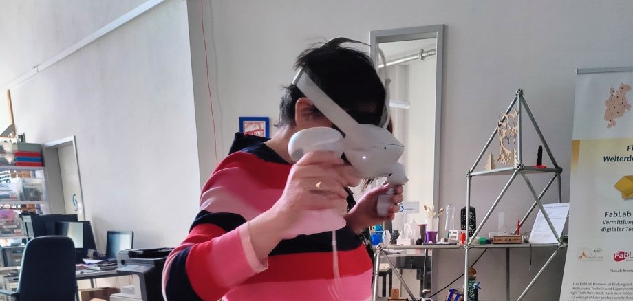 ZWAR-Bericht Person steht in einem offenen Kreativ- und Technikraum und nutzt eine Virtual-Reality-Brille mit zwei Controllern in den Händen. Im Hintergrund sind Arbeitsplätze, technische Geräte und Ausstellungsobjekte zu sehen.