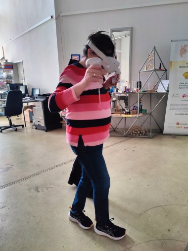ZWAR-Bericht Person steht in einem offenen Kreativ- und Technikraum und nutzt eine Virtual-Reality-Brille mit zwei Controllern in den Händen. Im Hintergrund sind Arbeitsplätze, technische Geräte und Ausstellungsobjekte zu sehen.