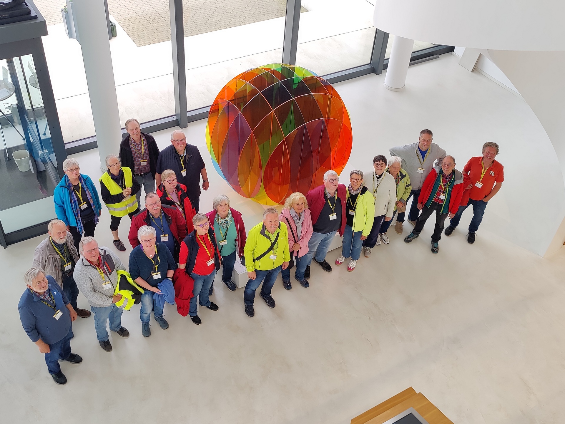 ZWAR bei Harting Gruppe von etwa 20 Menschen steht in einem hellen modernen Gebäude um eine große bunte Kunstinstallation aus transparenten farbigen Elementen. Die Personen schauen nach oben zur Kamera, einige tragen Namensschilder und bunte Jacken.