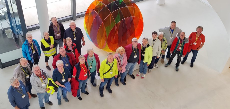 ZWAR bei Harting Gruppe von etwa 20 Menschen steht in einem hellen modernen Gebäude um eine große bunte Kunstinstallation aus transparenten farbigen Elementen. Die Personen schauen nach oben zur Kamera, einige tragen Namensschilder und bunte Jacken.