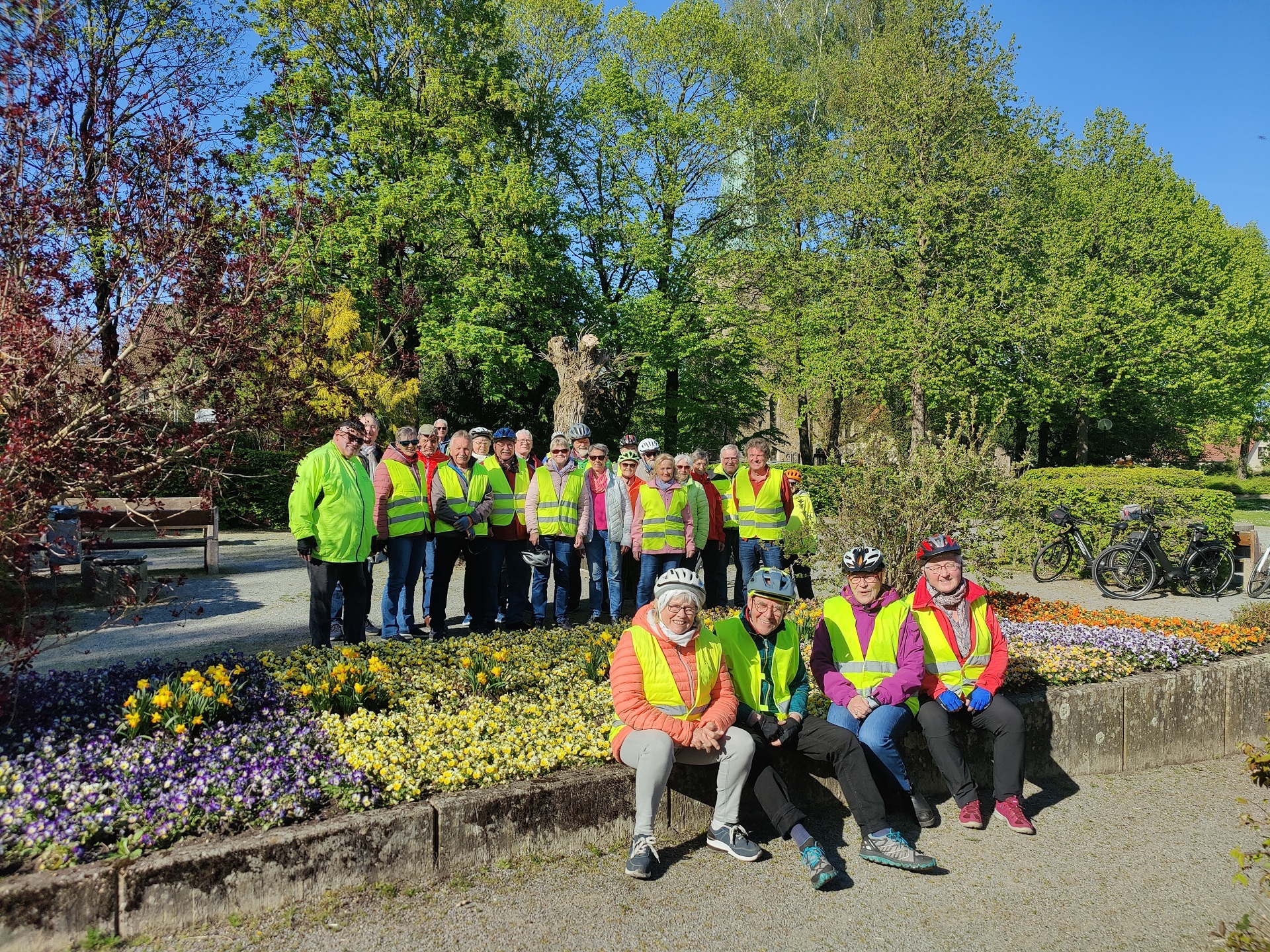 ZWAR-Tour Schledehausen Gruppe von Radfahrenden mit gelben Warnwesten steht und sitzt in einer Parkanlage mit bunten Blumenbeeten und hohen Bäumen. Einige tragen Fahrradhelme, im Hintergrund stehen mehrere Fahrräder. Die Szene wirkt sonnig und gemeinschaftlich.