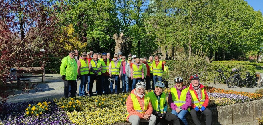ZWAR-Tour Schledehausen Gruppe von Radfahrenden mit gelben Warnwesten steht und sitzt in einer Parkanlage mit bunten Blumenbeeten und hohen Bäumen. Einige tragen Fahrradhelme, im Hintergrund stehen mehrere Fahrräder. Die Szene wirkt sonnig und gemeinschaftlich.